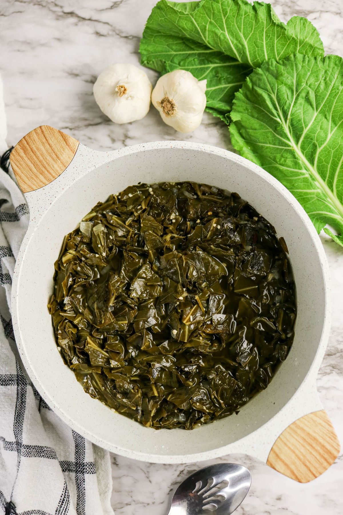 Cooked collard greens in a white pot, placed on a marble surface with fresh collard leaves, garlic bulbs, and a striped towel in the background. A spoon is nearby.