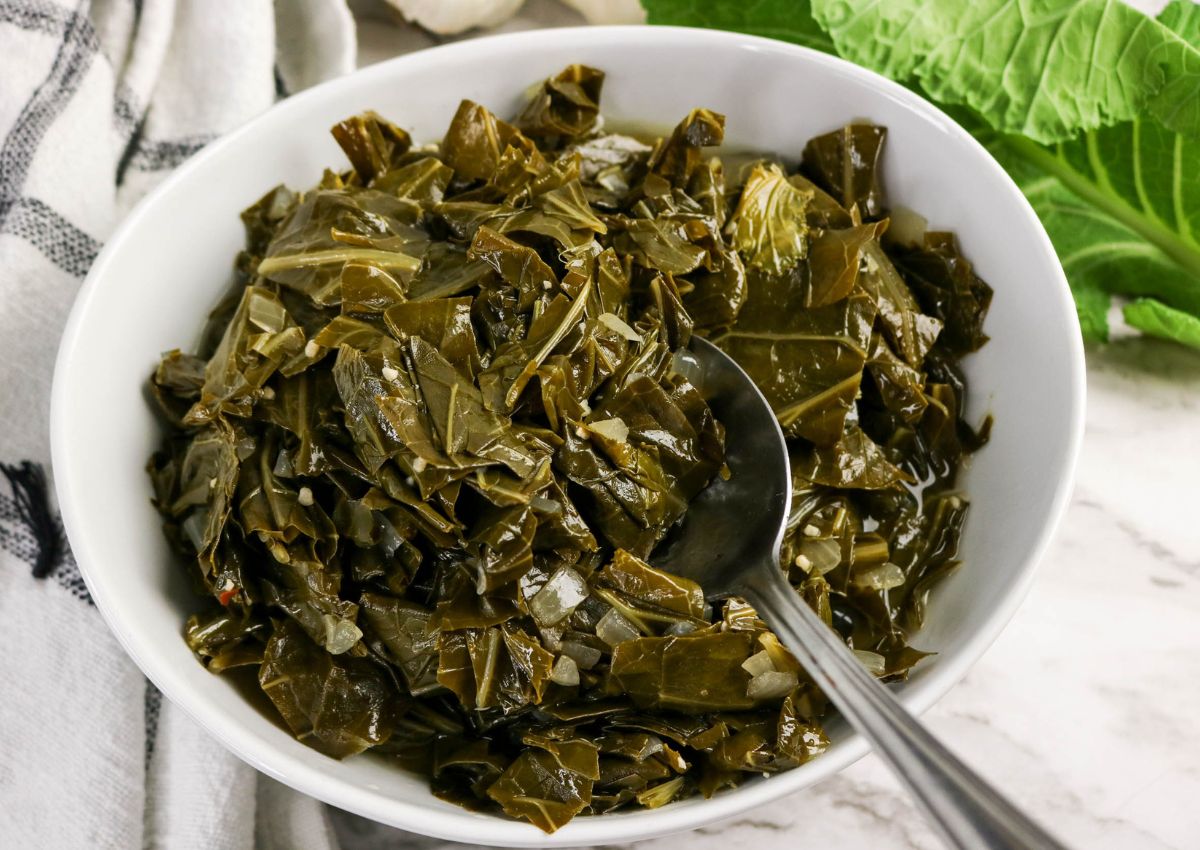 A white bowl filled with cooked collard greens, with a metal spoon resting inside; leafy greens and a towel are visible in the background.