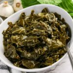 A white bowl filled with cooked collard greens, with a spoon resting inside, set on a marble surface with garlic and leafy greens in the background.