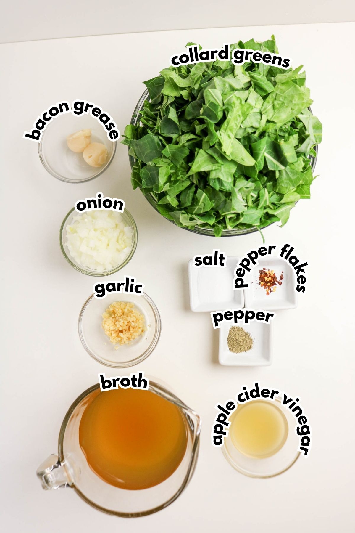 Overhead view of ingredients for cooking collard greens, including chopped collard greens, onion, garlic, bacon grease, broth, apple cider vinegar, salt, pepper, and pepper flakes.
