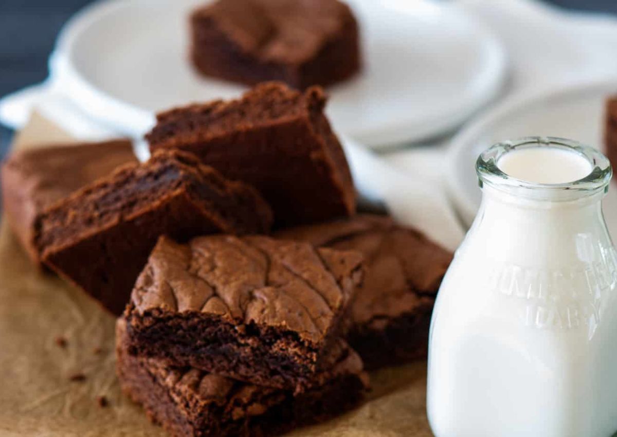 A stack of chocolate brownies on parchment paper next to a small glass bottle of milk, with plates and a brownie in the background.