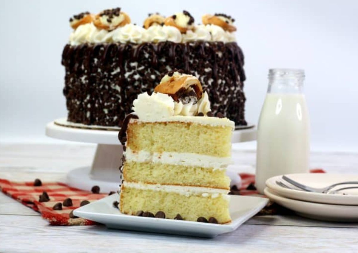 A slice of layered vanilla cake with cream and chocolate chips sits on a plate, in front of a whole decorated cake and a bottle of milk.