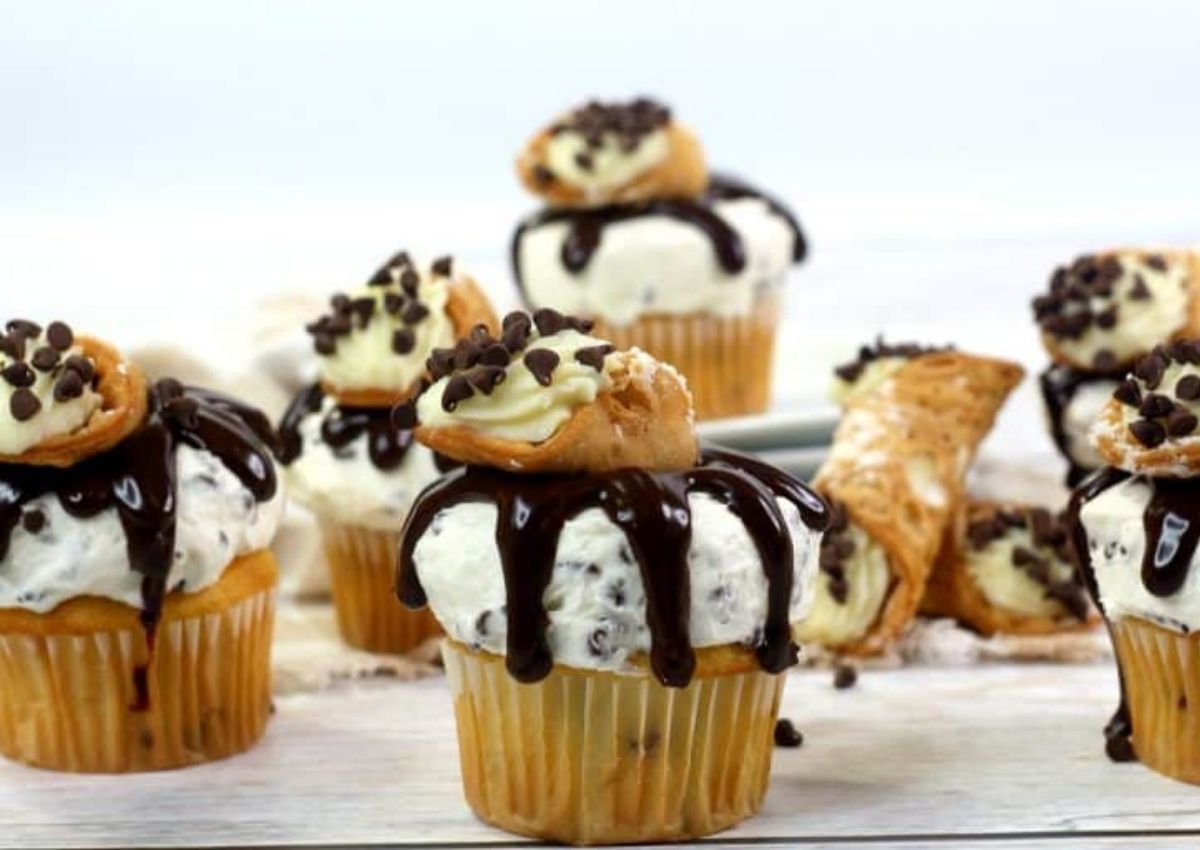 Cupcakes topped with whipped cream, chocolate chips, chocolate drizzle, and mini cannoli are arranged on a white surface.