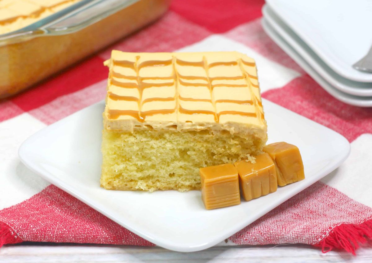 A square slice of yellow cake with caramel frosting sits on a white plate next to three caramel candies, on a red and white checkered cloth.