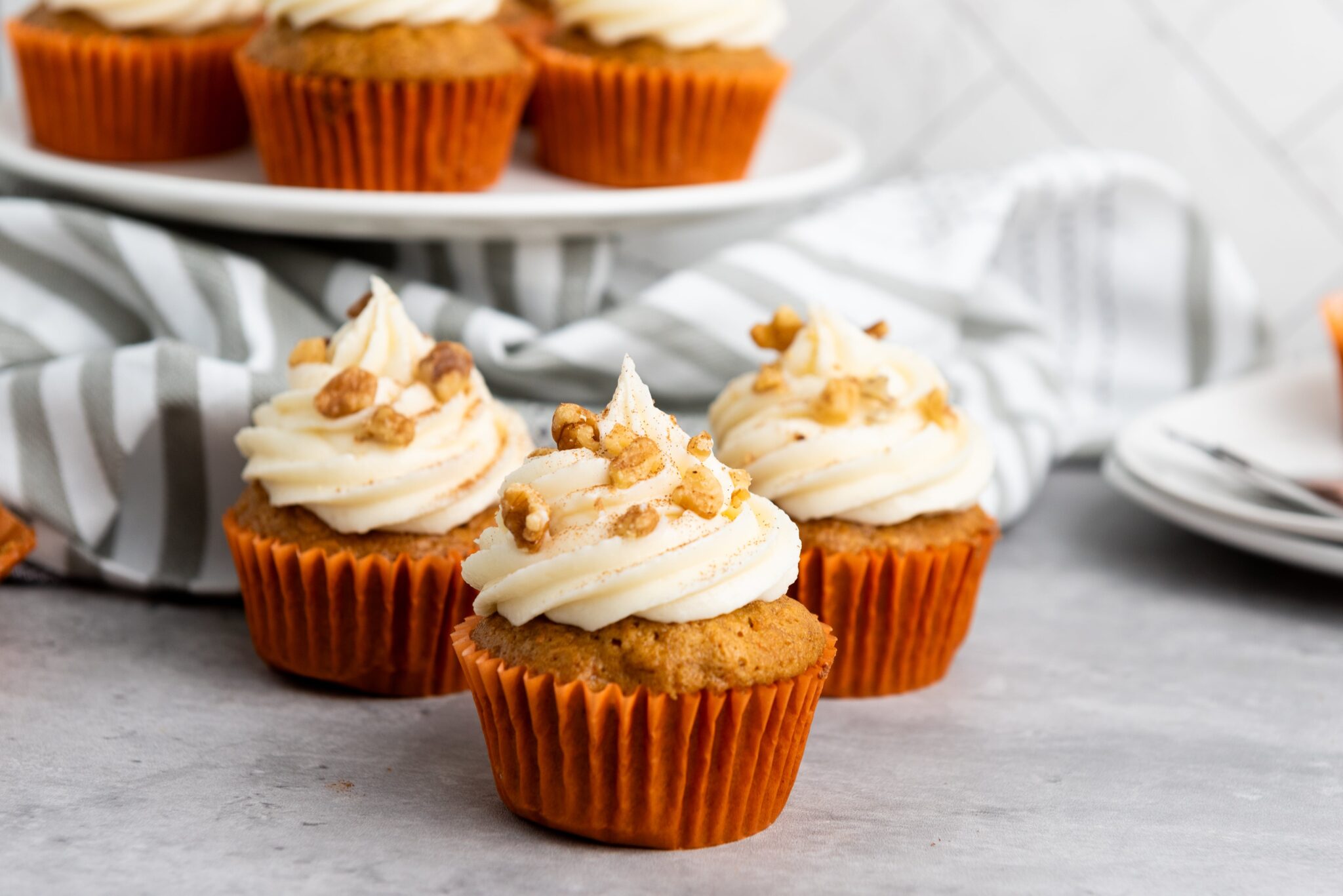 Three frosted cupcakes topped with chopped walnuts are arranged on a gray surface, with more cupcakes on a white plate in the background.