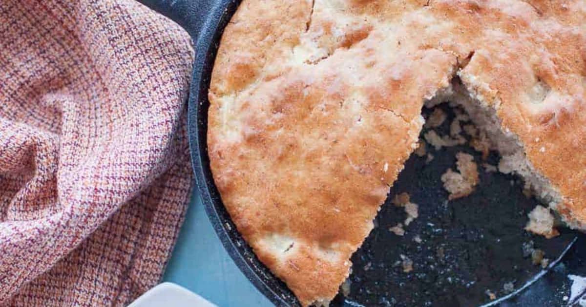 A round skillet with a baked golden-brown bread or cake, one wedge removed, next to a textured pinkish-brown cloth.