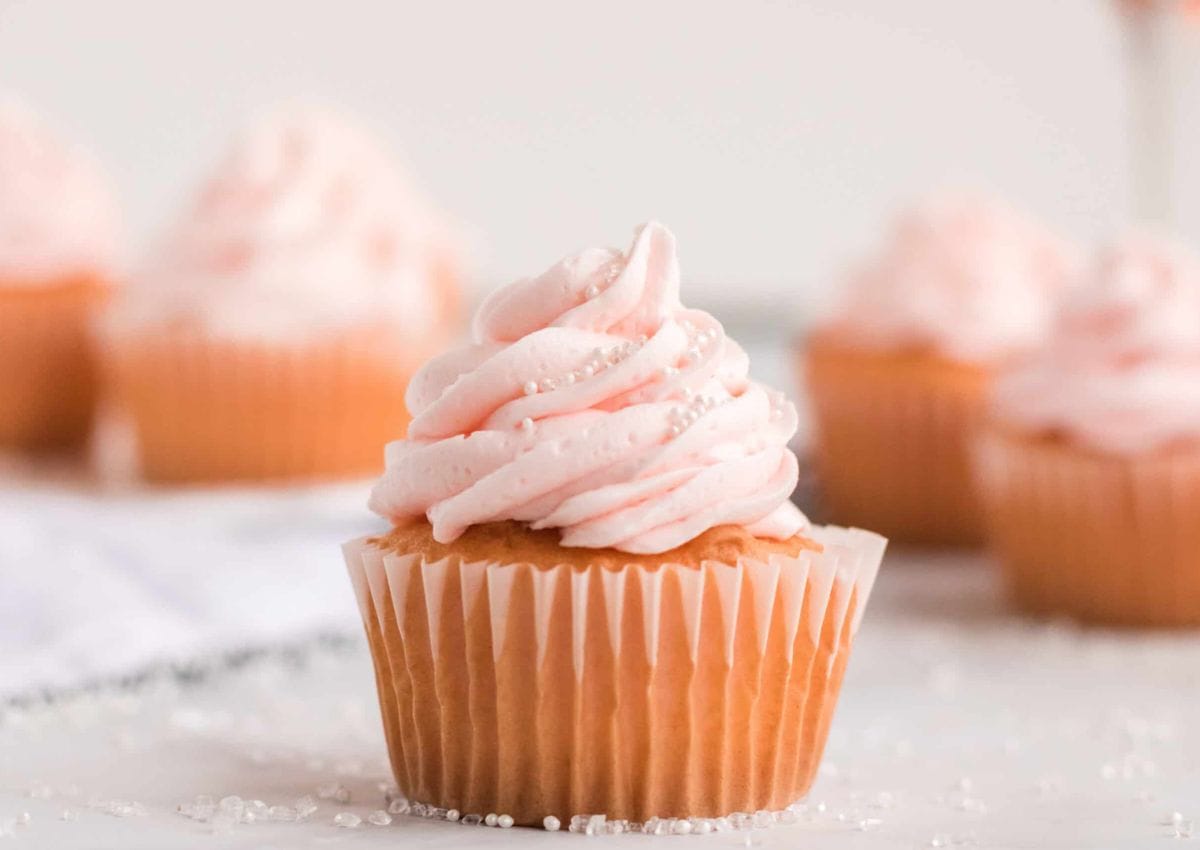 A cupcake with pink frosting and white sprinkles sits on a white surface, with more cupcakes blurred in the background.