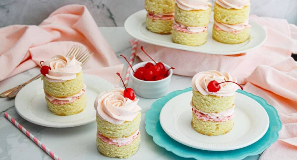 Individual round vanilla cakes stacked with pink frosting and topped with a cherry, served on plates with a bowl of cherries and pink napkins in the background.