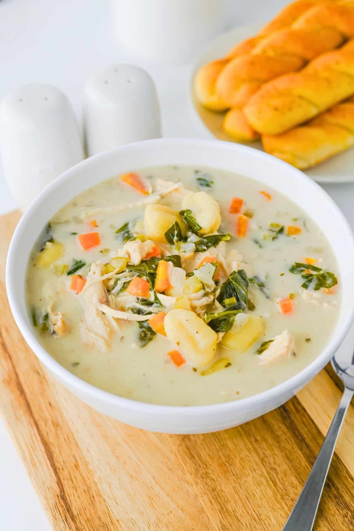 A white bowl of creamy chicken and vegetable soup with gnocchi sits on a wooden board, accompanied by breadsticks and salt and pepper shakers in the background.
