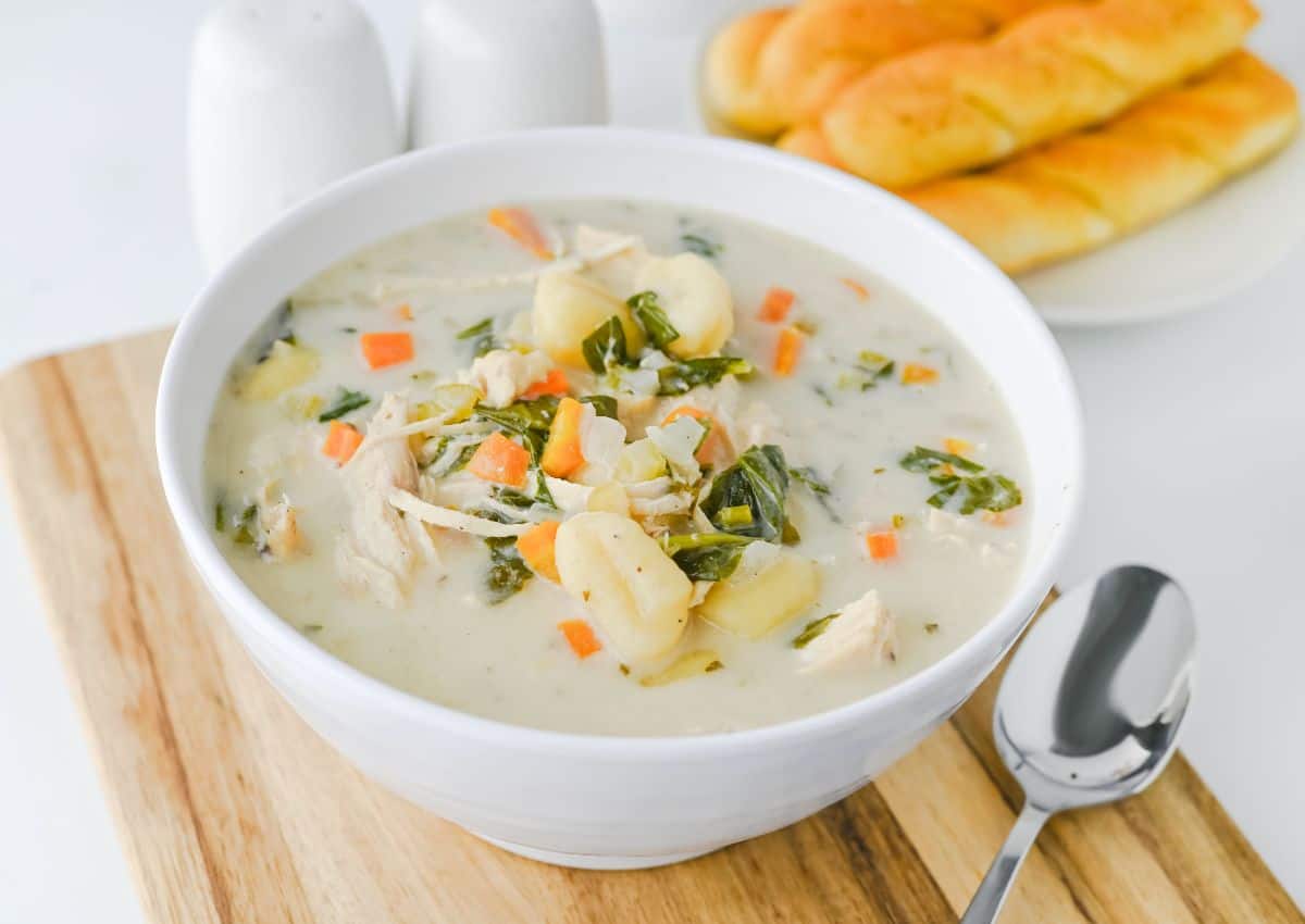 A bowl of creamy chicken and vegetable soup with gnocchi, served on a wooden board next to a spoon and a plate of breadsticks in the background.
