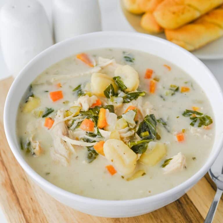 A bowl of creamy chicken and vegetable soup with gnocchi, carrots, spinach, and potatoes, served on a wooden board with breadsticks in the background.