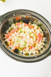 Chopped onions, celery, and carrots in a black slow cooker on a white surface.