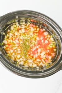 A slow cooker filled with chopped vegetables, broth, and herbs, ready to be cooked.