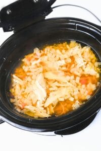 A slow cooker filled with shredded chicken, diced onions, and carrots in broth, with the lid open.
