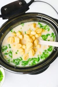 A slow cooker filled with a creamy soup containing gnocchi, chopped greens, and a white spatula.