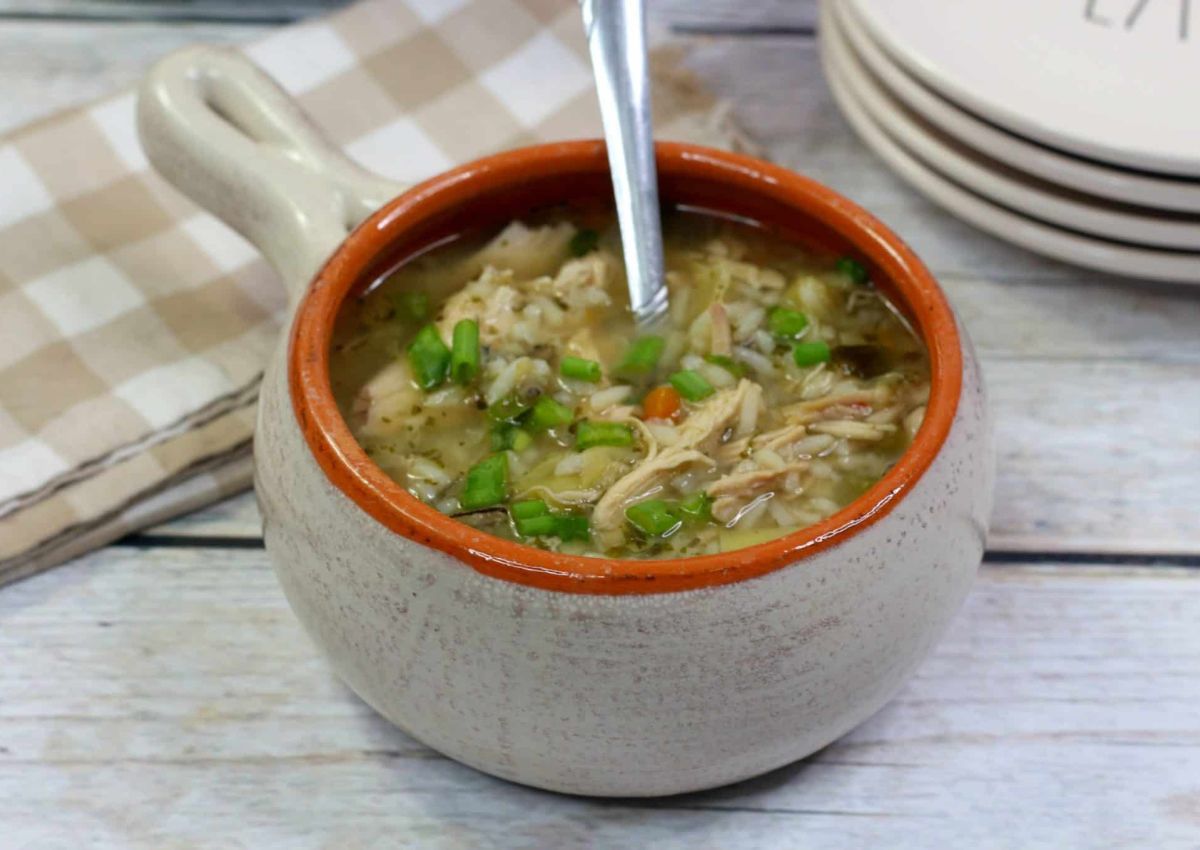 A ceramic bowl filled with chicken and rice soup, garnished with chopped green onions, with a spoon inside. A checked napkin and stacked plates are in the background.