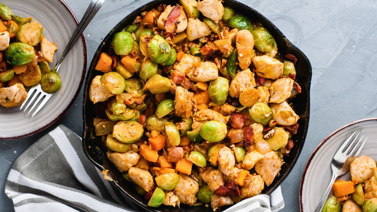 A skillet filled with cooked Brussels sprouts, diced chicken, bacon, and sweet potatoes, with portions served on two plates nearby.