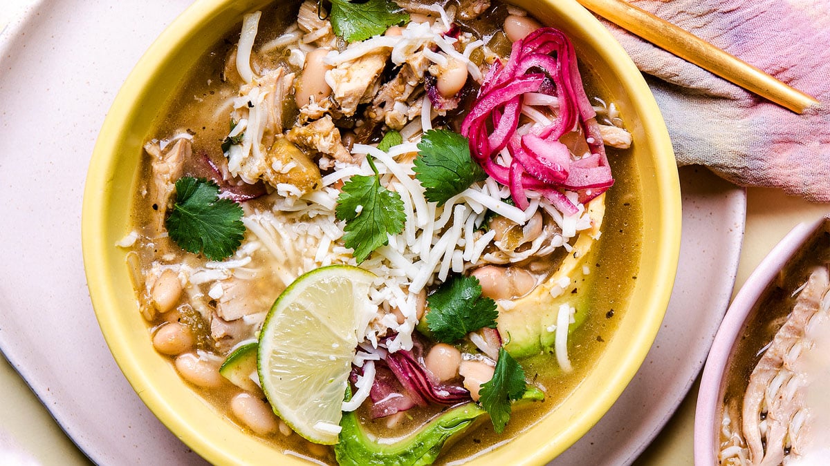 A bowl of chili with shredded cheese, white beans, cilantro, pickled onions, avocado slices, and a lime wedge on top.