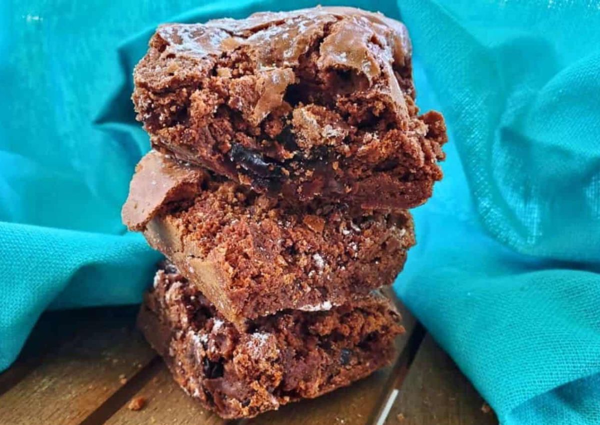 Three chocolate brownies are stacked on top of each other, with a blue cloth in the background and a wooden surface beneath them.