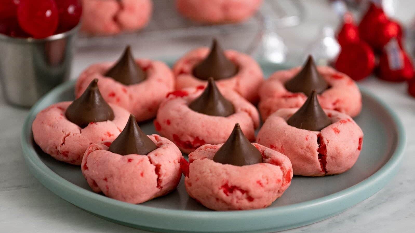 A plate of pink cherry cookies topped with chocolate kisses, with more cookies and cherries in the background.