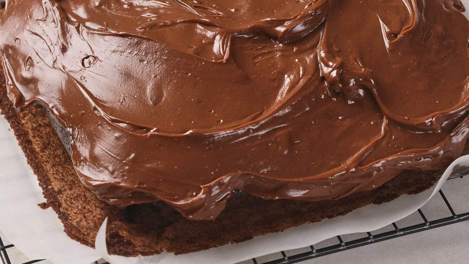 A square chocolate cake with thick chocolate frosting sits on a cooling rack lined with parchment paper.