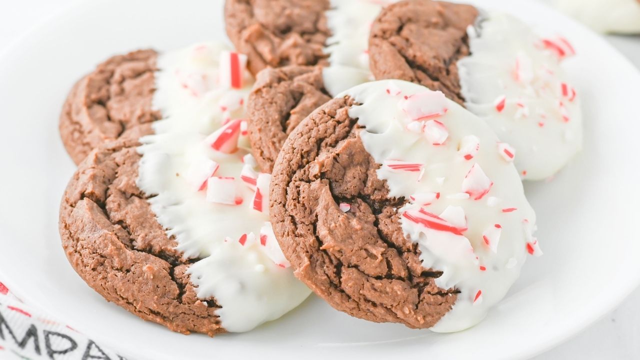 Chocolate cookies partially dipped in white chocolate and sprinkled with crushed peppermint pieces, arranged on a white plate.