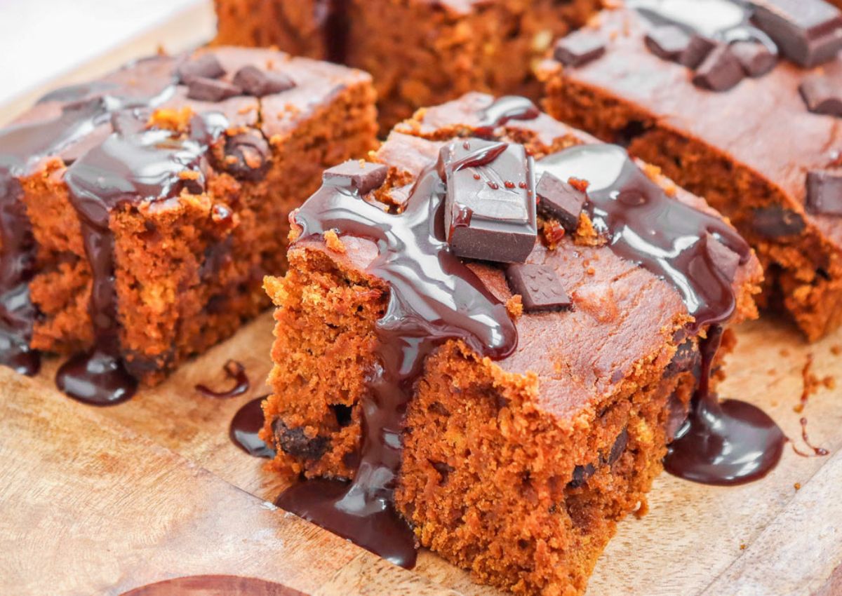 Squares of chocolate cake topped with chocolate chunks and drizzled with chocolate sauce, arranged on a wooden board.