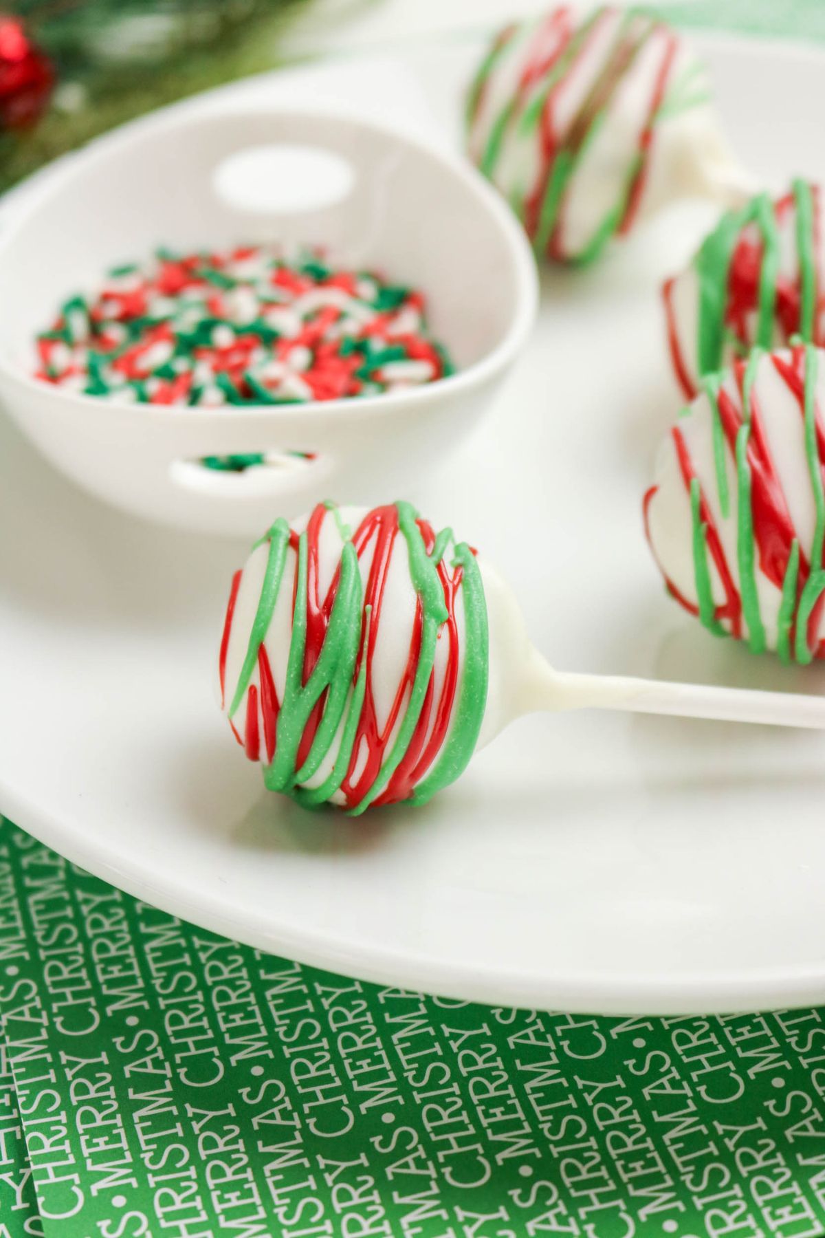 Round cake pops covered in white chocolate with red and green drizzle, displayed on a white plate next to a small bowl of festive sprinkles.