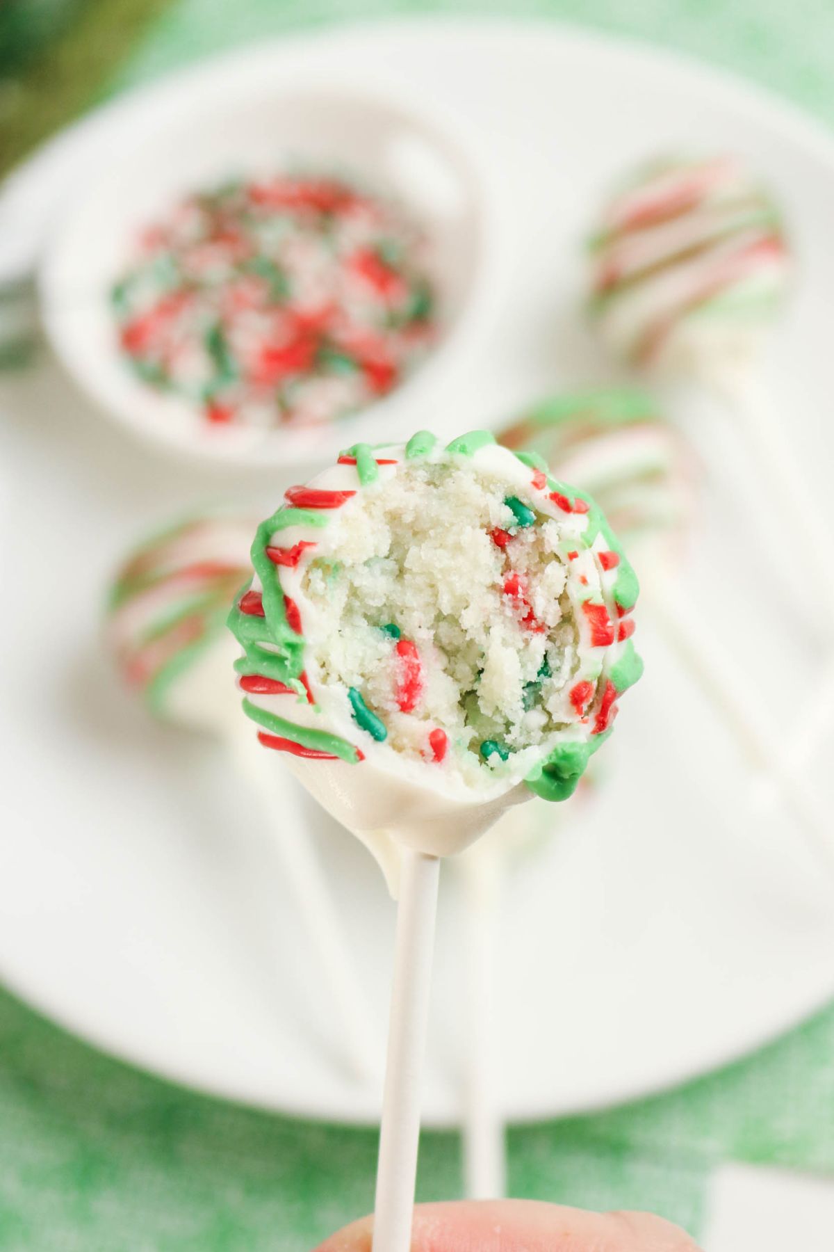 A close-up of a cake pop with white icing and red and green drizzle, showing a bite taken out to reveal a colorful, sprinkled interior. More cake pops are on a white plate in the background.