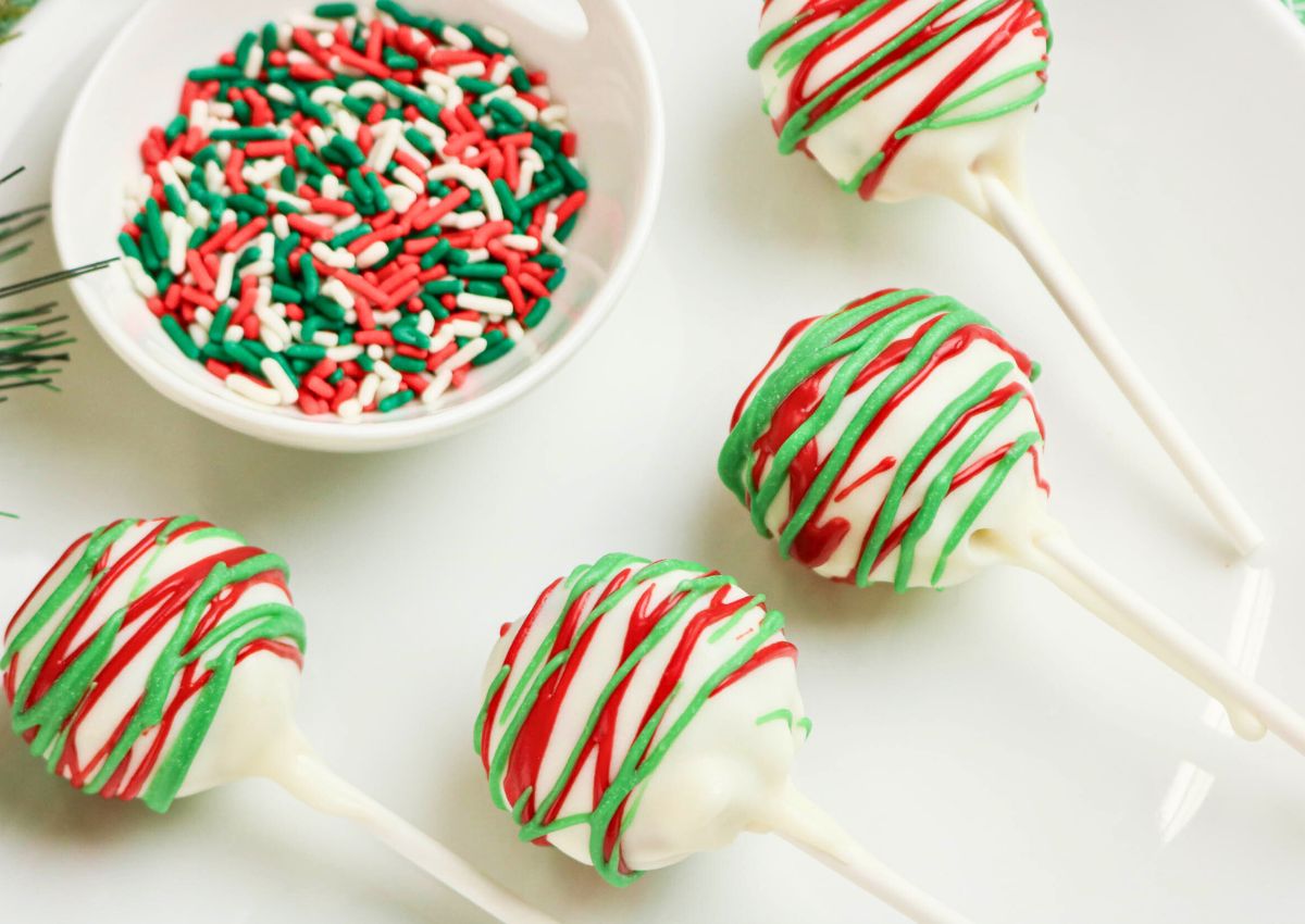 Four white chocolate cake pops with red and green drizzle are arranged beside a bowl of red, green, and white sprinkles on a white plate.