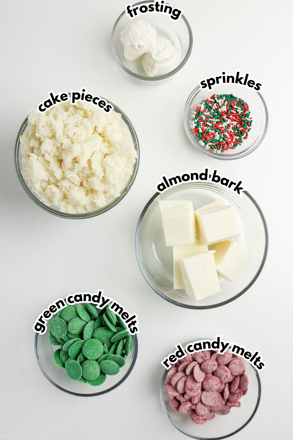 Six bowls filled with cake pieces, frosting, sprinkles, almond bark, green candy melts, and red candy melts are arranged on a white surface, each labeled with text.
