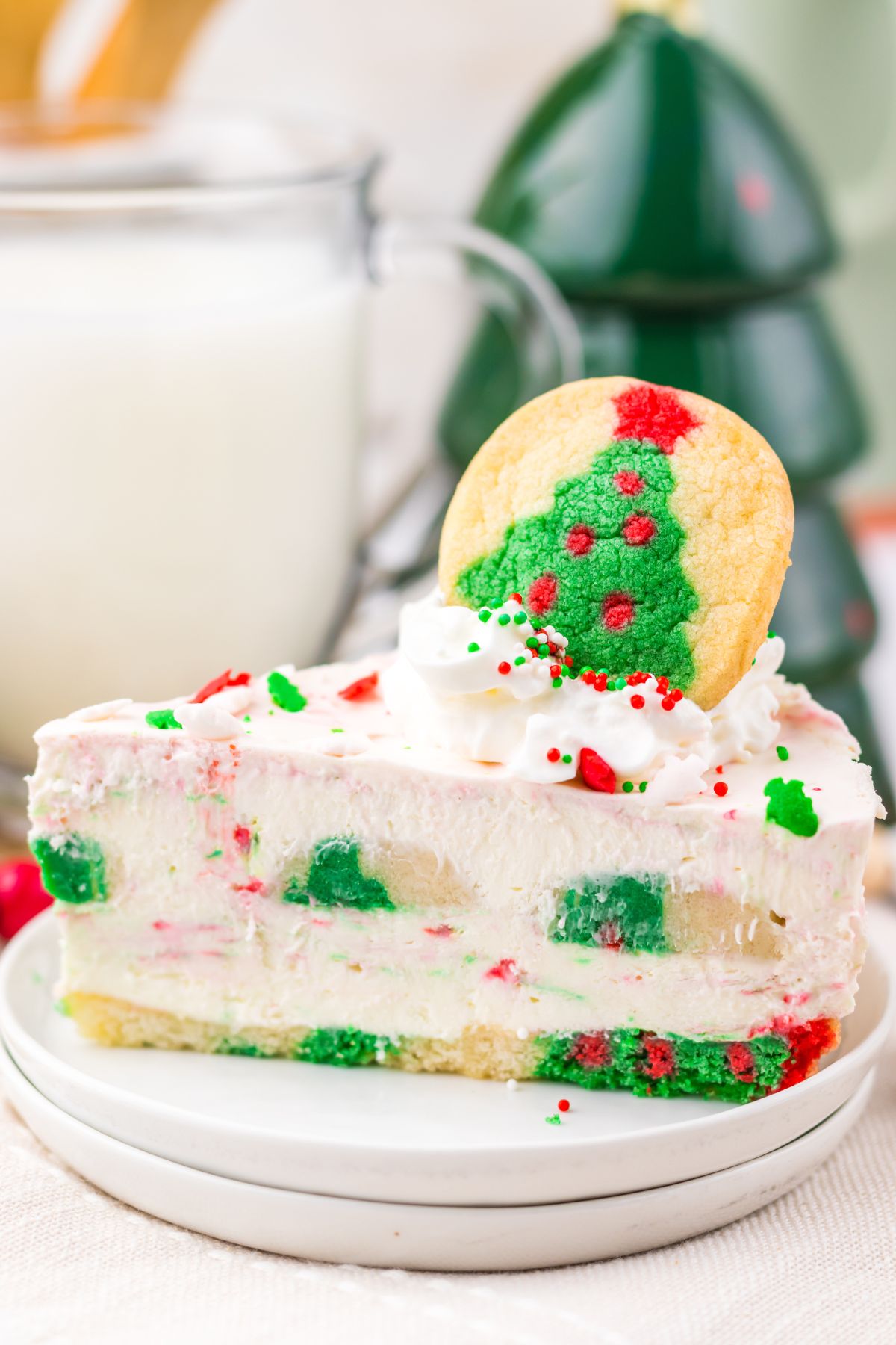 A slice of festive cake with red and green sprinkles, topped with whipped cream and a Christmas tree cookie, served on a white plate.