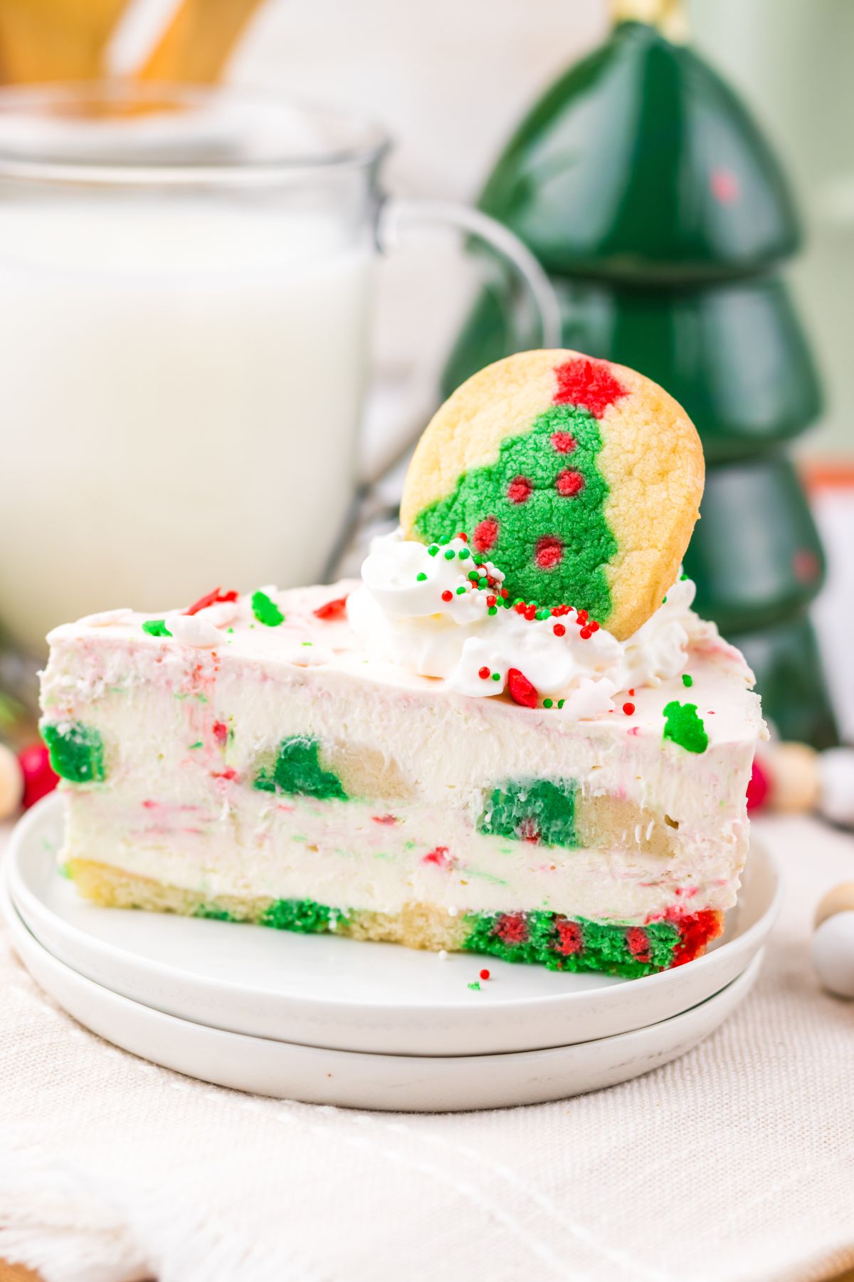 A slice of festive cheesecake with red and green sprinkles, topped with whipped cream and a Christmas tree cookie, sits on a small white plate.