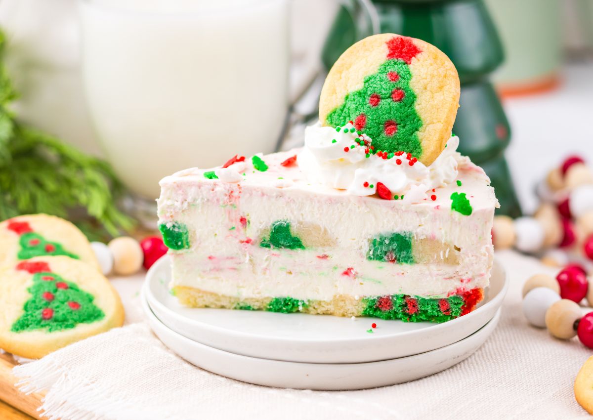 A slice of festive cheesecake topped with whipped cream, sprinkles, and a Christmas tree sugar cookie, served on a white plate.