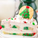 A slice of festive cheesecake with red and green sprinkles, topped with whipped cream and a Christmas tree-shaped cookie.