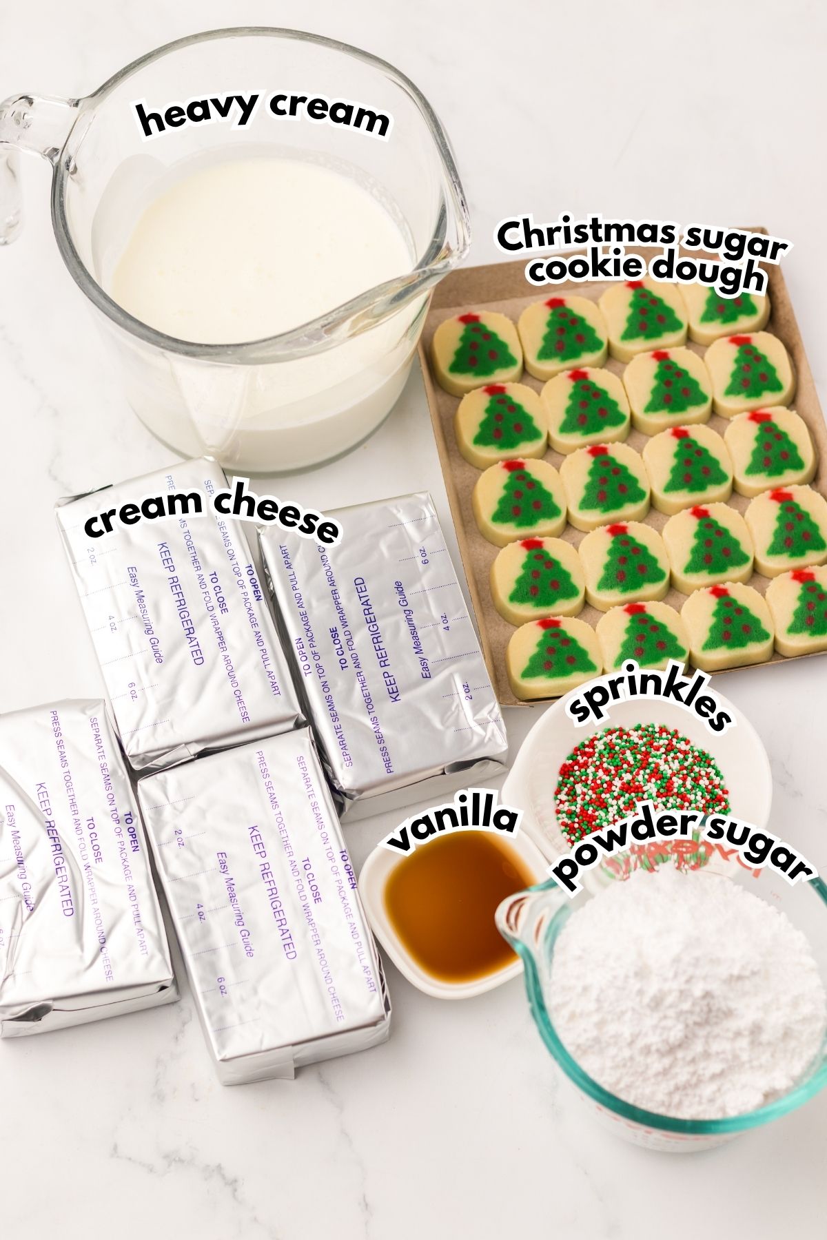 A glass measuring cup of heavy cream, four blocks of cream cheese, a bowl of powdered sugar, a small bowl of vanilla, sprinkles, and Christmas tree sugar cookies are displayed on a counter.