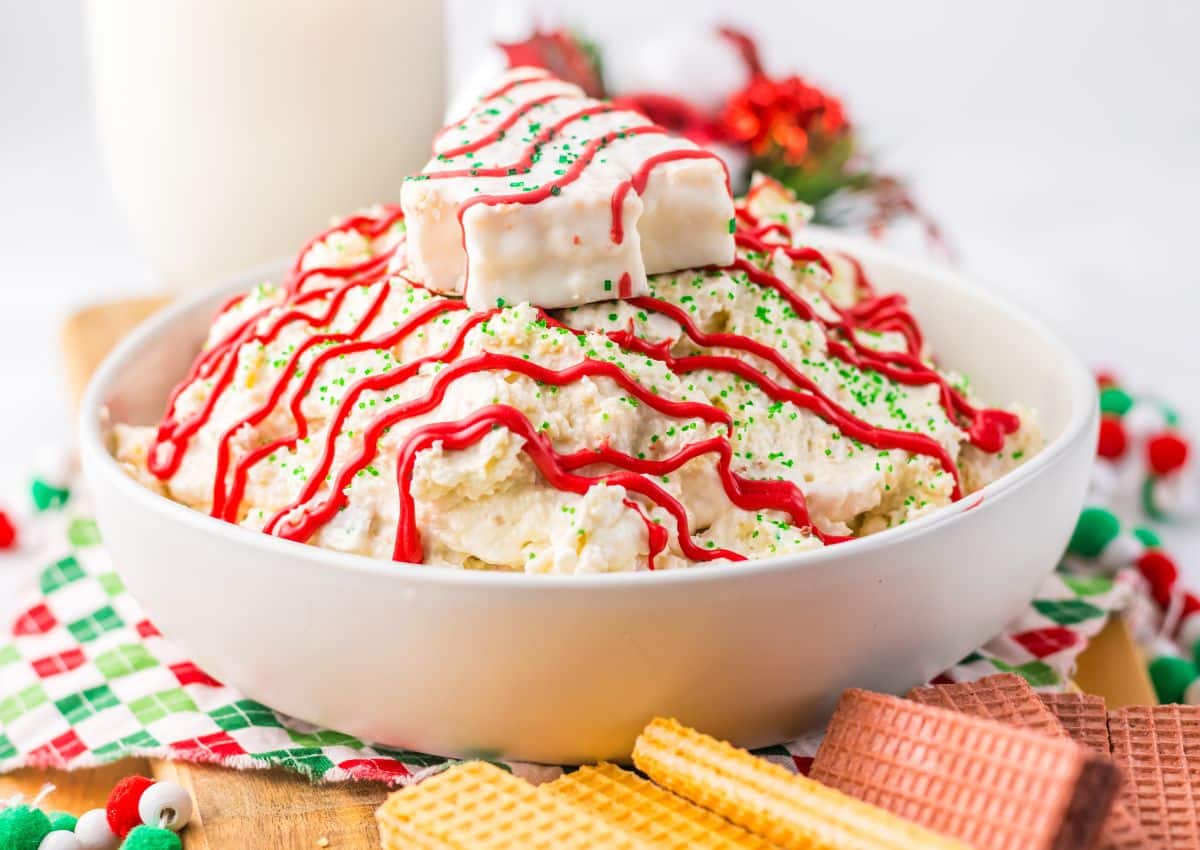 A bowl of creamy dip topped with red icing, green sprinkles, and a white-frosted snack cake, surrounded by colorful wafer cookies.