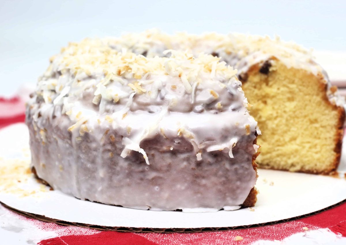 A round bundt cake with white icing and toasted coconut flakes on top, with one slice removed, displayed on a white surface.