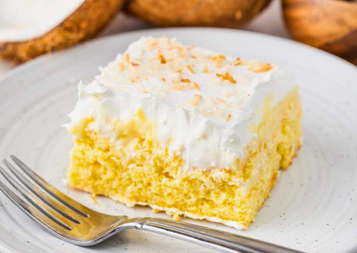 A slice of yellow cake with white frosting and toasted coconut flakes on a white plate, with a fork beside it.