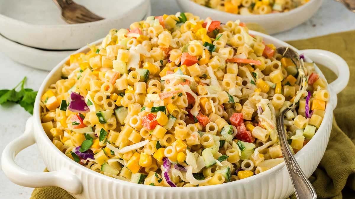 A large bowl of macaroni salad with chopped vegetables and herbs, mixed in a creamy dressing, with serving utensils placed inside.
