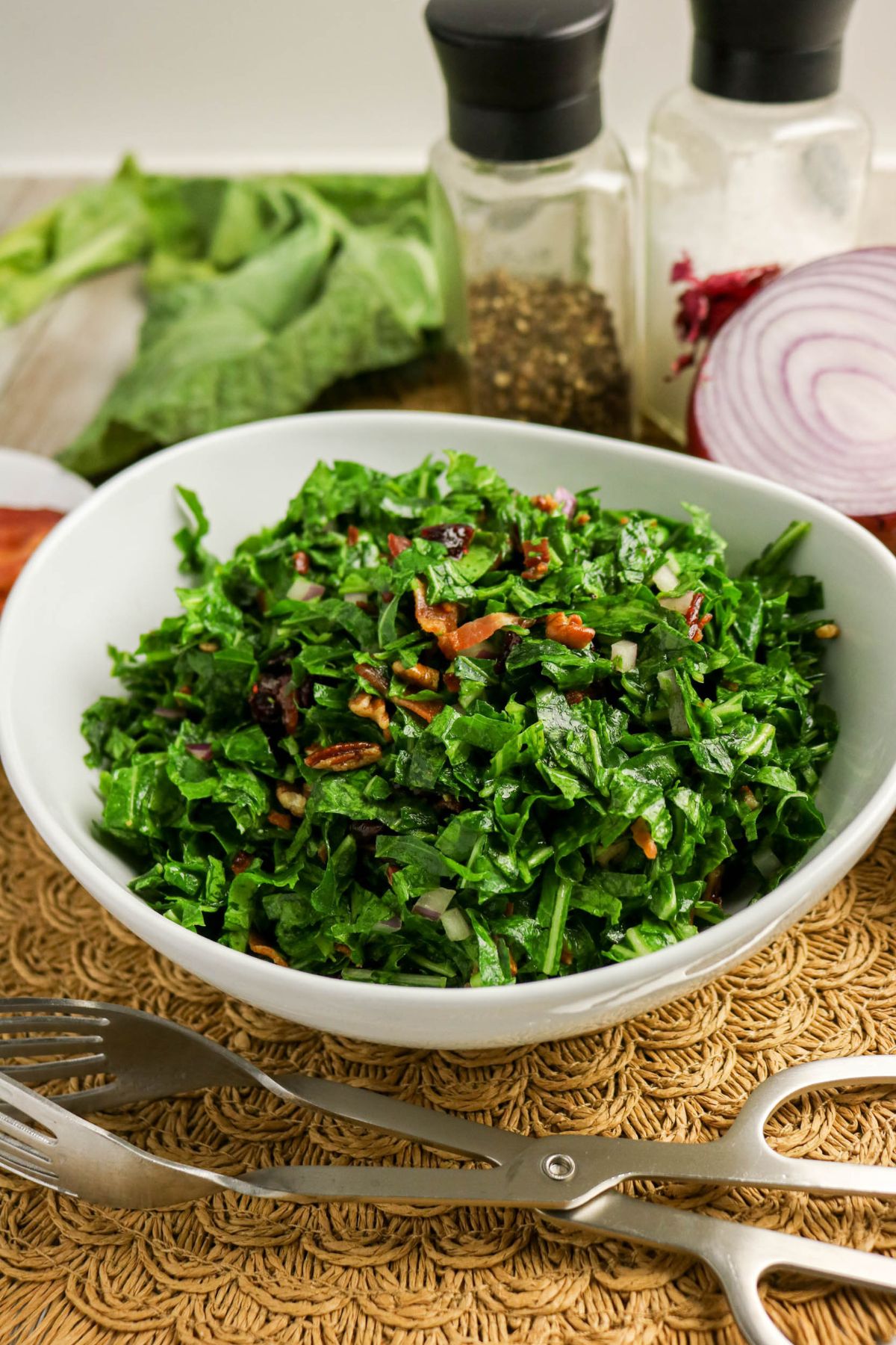 A bowl of chopped leafy green salad with bacon and onions on a woven mat, with salad tongs and seasonings in the background.