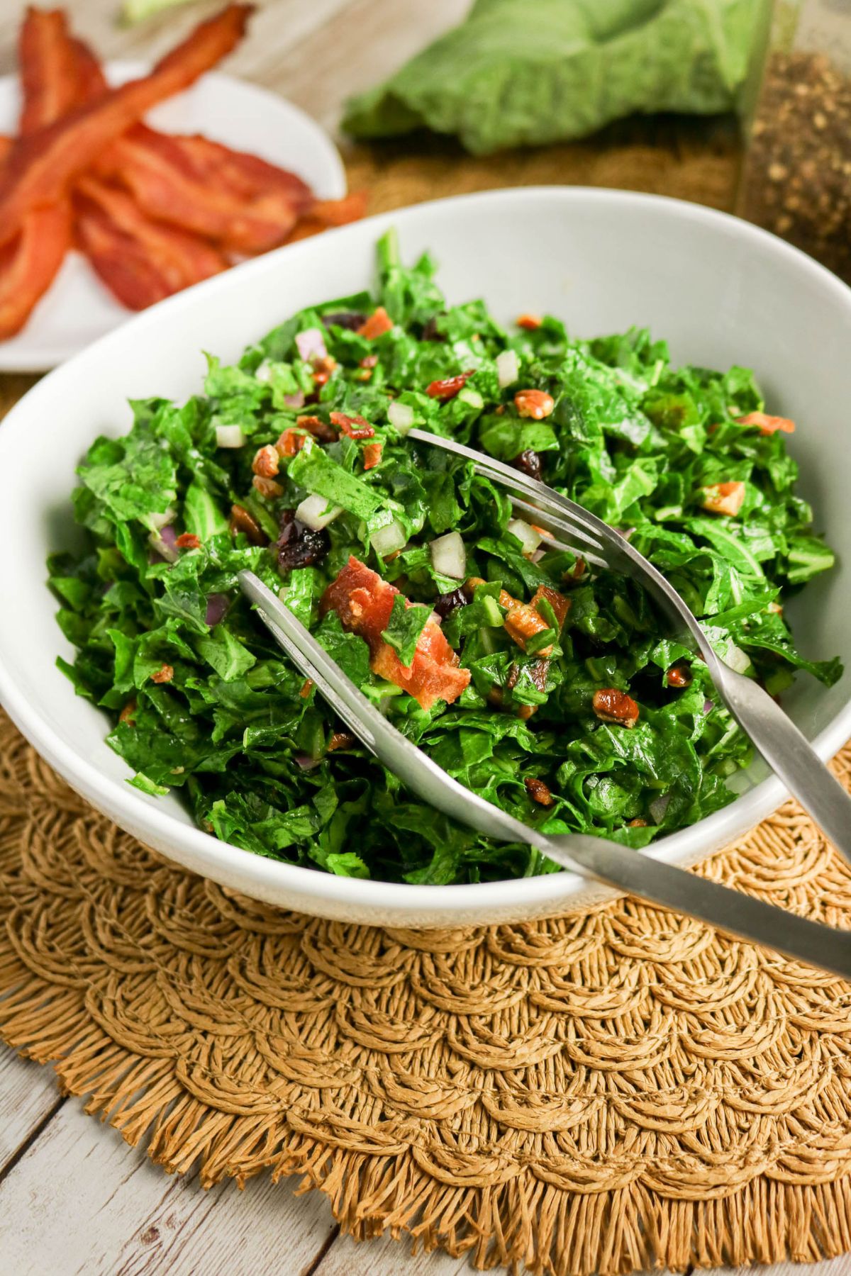 A white bowl filled with chopped kale salad, topped with bacon bits and diced vegetables, sits on a woven placemat with metal tongs resting inside.