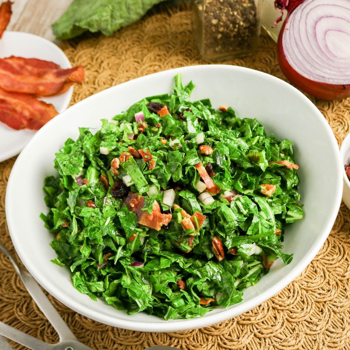A white bowl filled with chopped salad containing lettuce, bacon pieces, red onion, and dressing, placed on a woven placemat with salad ingredients nearby.