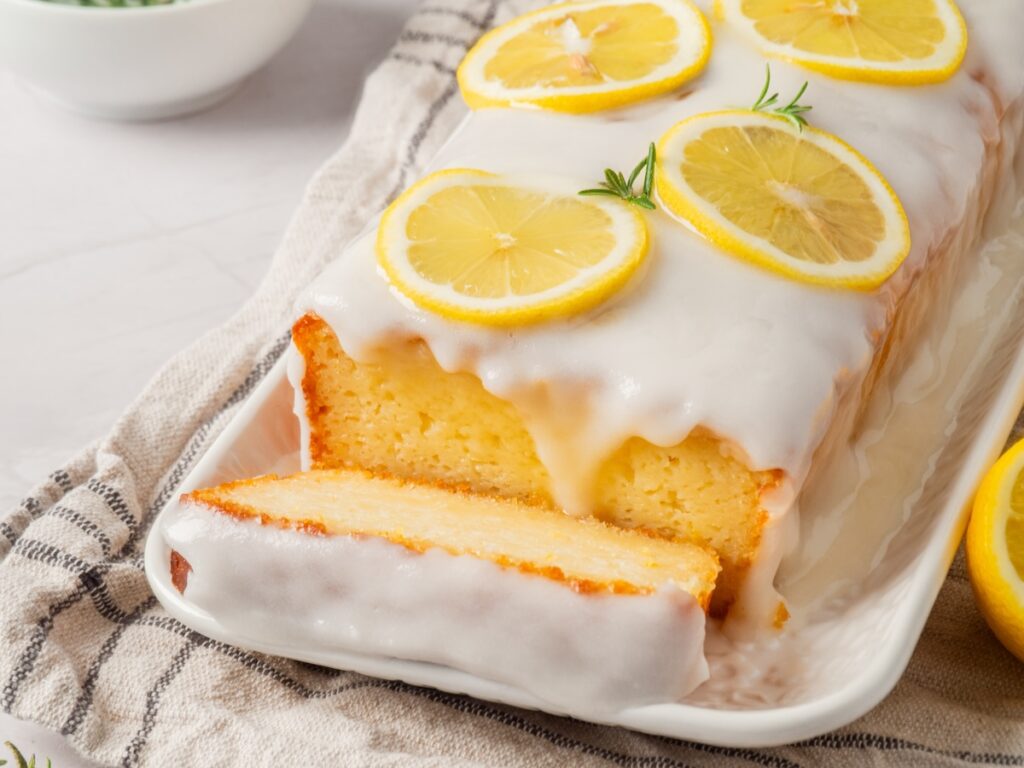 A loaf of lemon pound cake with white icing and lemon slices on top, placed on a white platter, with one slice cut.
