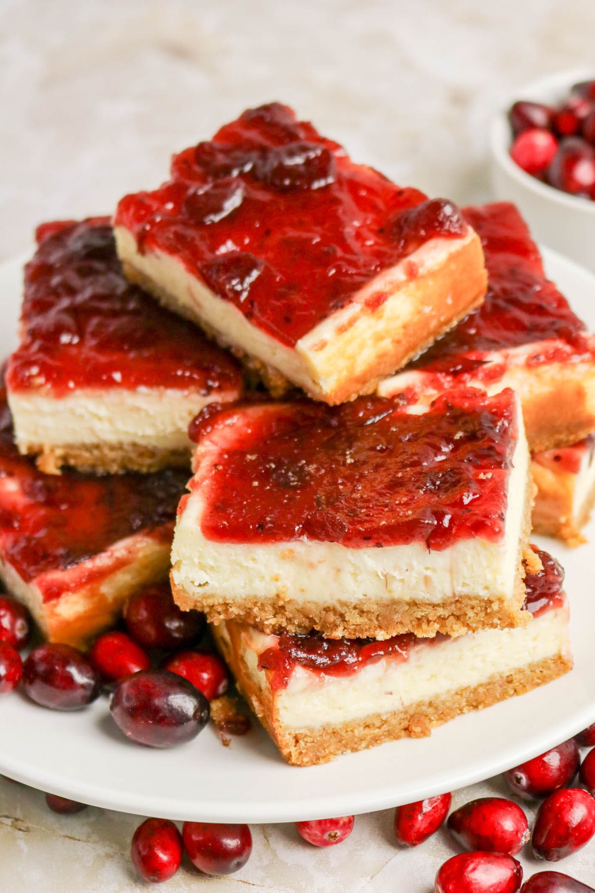 A plate of cheesecake bars with a golden crust and a glossy red cranberry topping, surrounded by fresh cranberries.