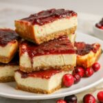 A plate of cheesecake bars with a graham cracker crust and cranberry topping, garnished with fresh cranberries.