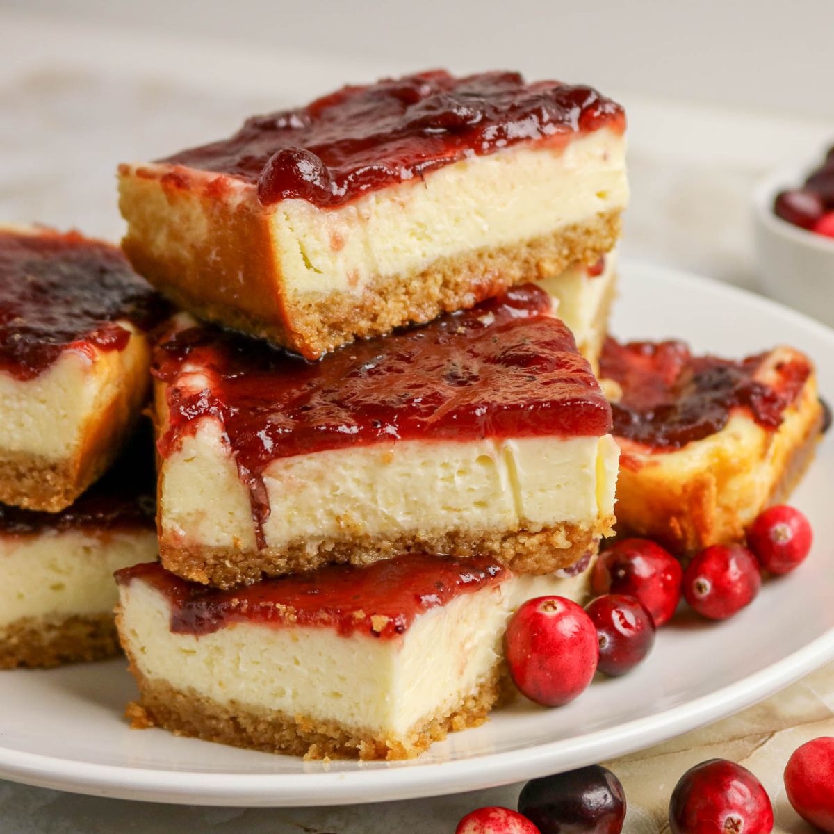 A plate of cheesecake bars with a graham cracker crust and cranberry topping, garnished with fresh cranberries.