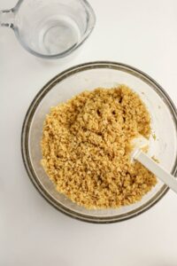 A glass bowl filled with a crumbly graham cracker mixture and a white spatula, next to an empty measuring cup on a white surface.