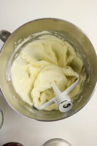 A mixing bowl with creamy, pale yellow frosting and a paddle attachment, shown from above on a white surface.