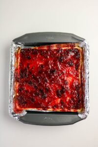 A square baking pan lined with foil containing a dessert topped with a layer of red fruit or berry preserves.